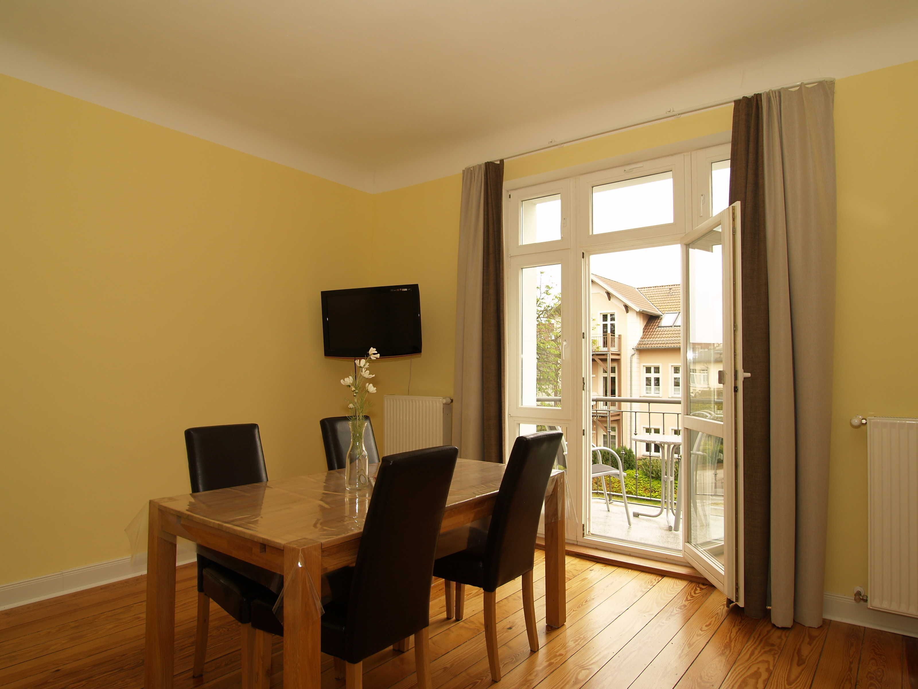 Ein Essbereich mit Holztisch und Stühlen, einer Blumenvase, einem Flachbildfernseher an der Wand und einer Glastür zum Außenbalkon.