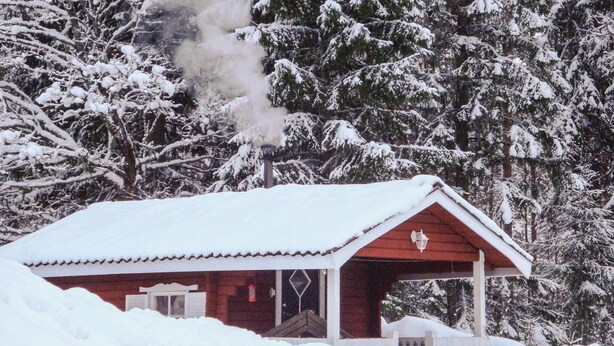 A snow-covered cabin with a chimney emitting smoke, surrounded by dense, snow-laden trees.