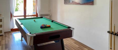 Una mesa de billar en una habitación con ventana, un cuadro en la pared y una puerta.