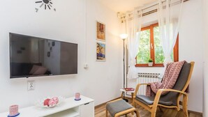 Un salon équipé d’un téléviseur à écran plat, d’un meuble de rangement blanc, d’une chaise en bois, d’une petite table et d’une fenêtre avec des rideaux.