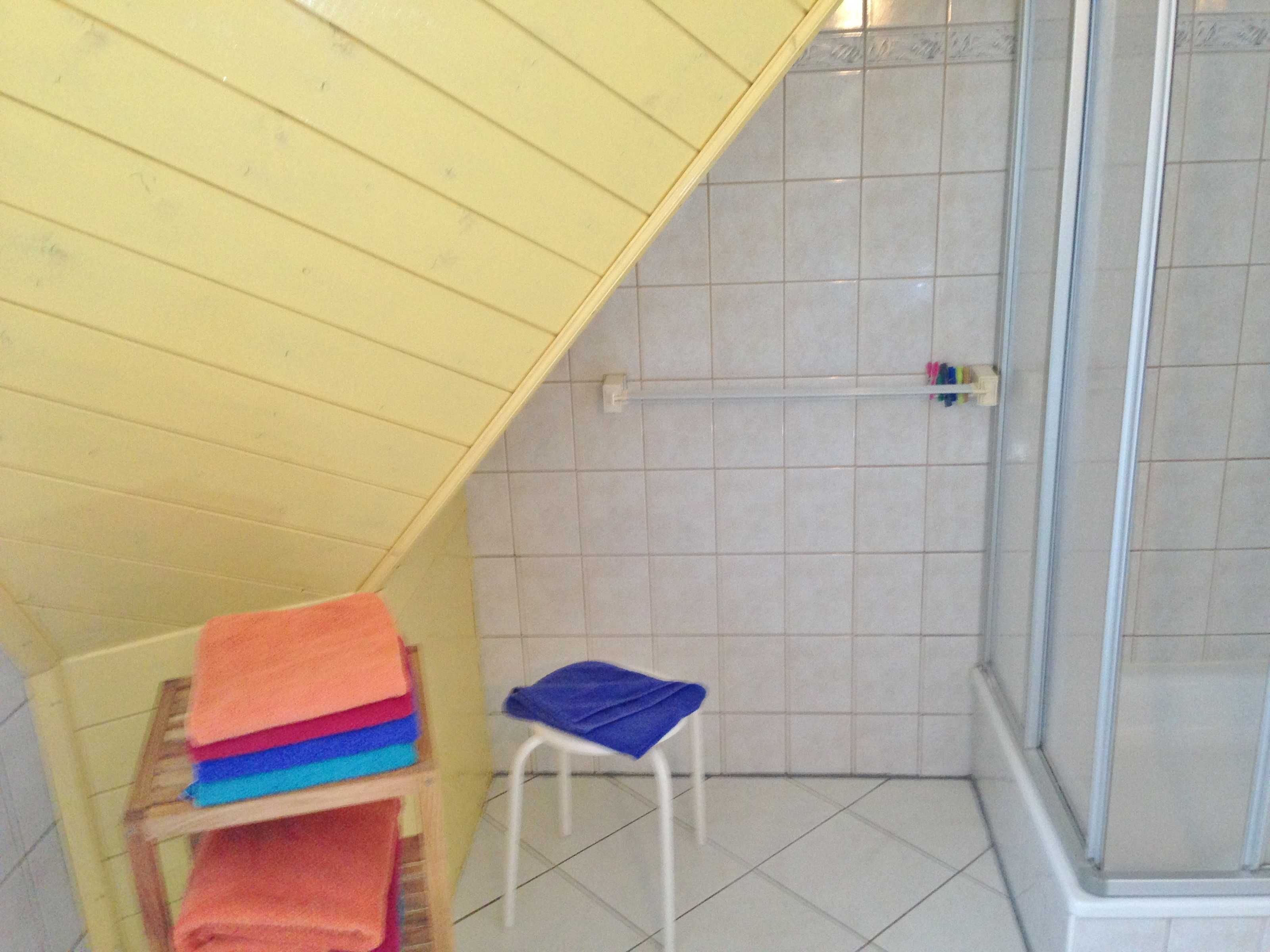 A small bathroom with a shower, a wooden shelf with towels, and a white chair.