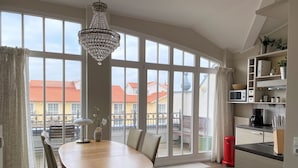 A dining area with a wooden table and chairs, a chandelier, and large windows with a view of buildings. - (76) 3-room apartment with roof terrace lake view - Appartementhaus Atlantik (Kühlungsborn)