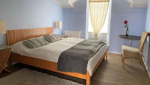 A bedroom with a bed, bedside tables, a chair, a small table with a bottle and a flower, and a window with curtains. - (76) 3-room apartment with roof terrace lake view - Appartementhaus Atlantik (Kühlungsborn)