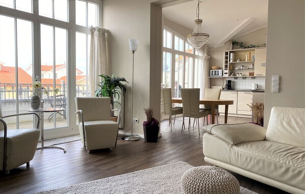A modern living room with a sofa, chairs, a dining table, and a kitchen area. - (76) 3-room apartment with roof terrace lake view - Appartementhaus Atlantik (Kühlungsborn)