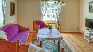 Una habitación con una cama de madera, un sofá morado, una silla de madera, una mesita con un jarrón, una televisión y una ventana con cortinas.