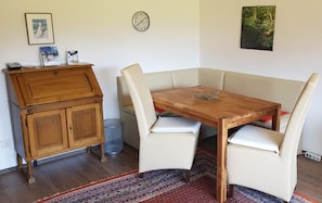 A dining area with a wooden table and four chairs, a clock on the wall, and a cabinet with a trash can.
