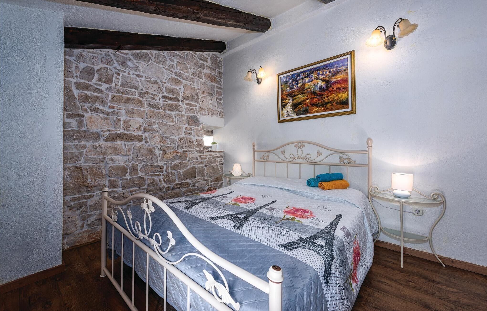 A bedroom with a stone wall, a bed with a blue quilt, a painting of a village, and two wall-mounted lamps.