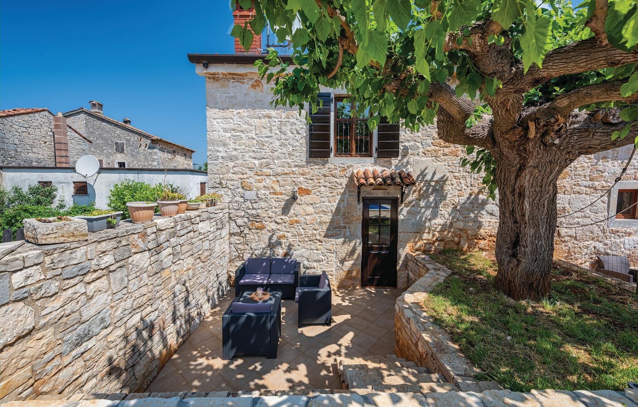 A stone building with a narrow courtyard, outdoor seating, and a large tree providing shade.