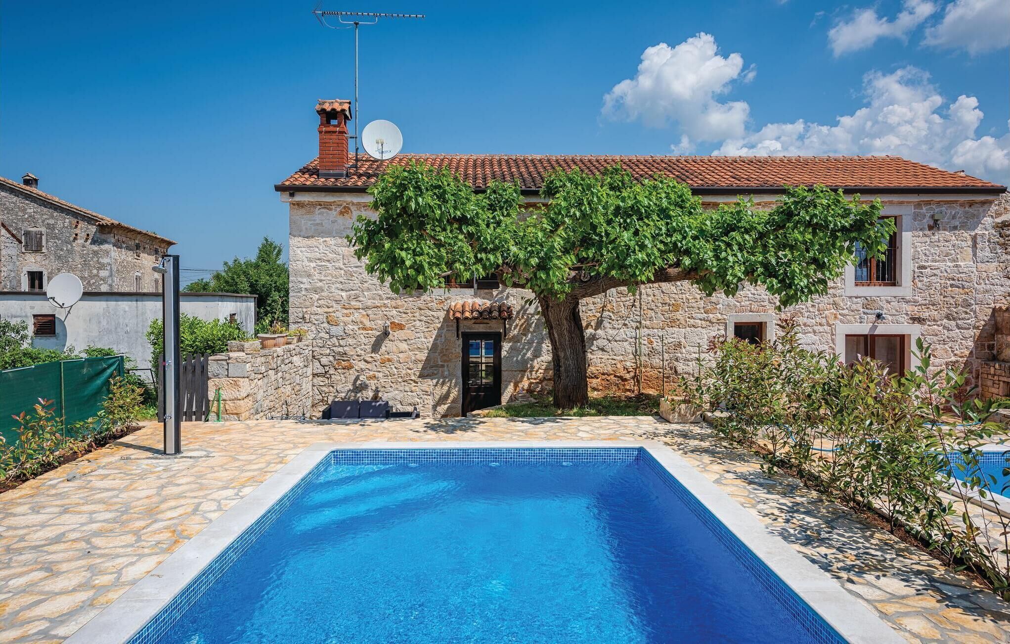Una villa in pietra con piscina, un albero di fronte e un cielo azzurro limpido.