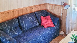 Habitación de esquina con pared de paneles de madera, un sofá azul estampado con un cojín rojo, una lámpara de pie y un televisor montado encima del sofá.