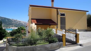 A yellow building with a red-tiled roof, a gated entrance, and a view of a blue body of water and mountains in the background. - Holiday apartment with terrace (Baška)