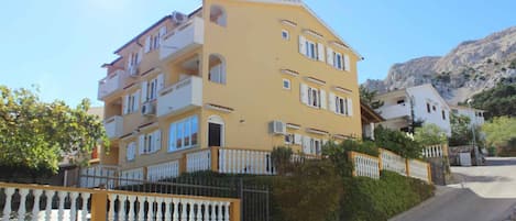 A multi-story yellow building with balconies, a gated entrance, and a mountainous backdrop.