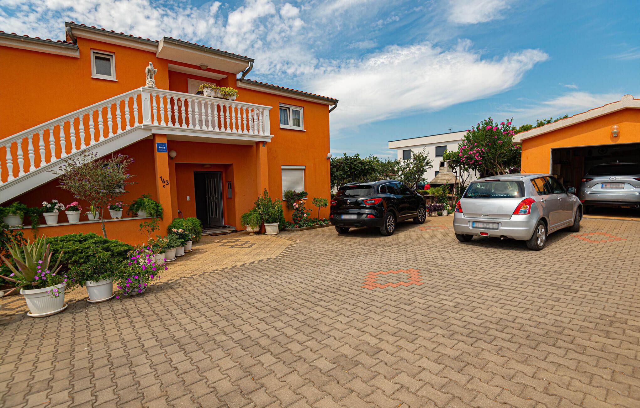 A building with a red facade, a white balcony, and a paved driveway with two cars parked.