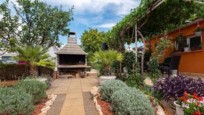 An outdoor patio with a stone fireplace, surrounded by various plants and a pergola with hanging plants. - 1 bedroom beautiful apartment in Vodice (Vodice)