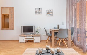 Ett modernt vardagsrum med ett skrivbord i trä, en platt-TV, en matplats med stolar och en prydnadsväxt.