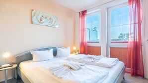 A neatly made bed with white linens and towels, a bedside lamp, a small table with a lamp, and a window with red curtains. - Dune Residence B 05 - DR B 05 (Heringsdorf)
