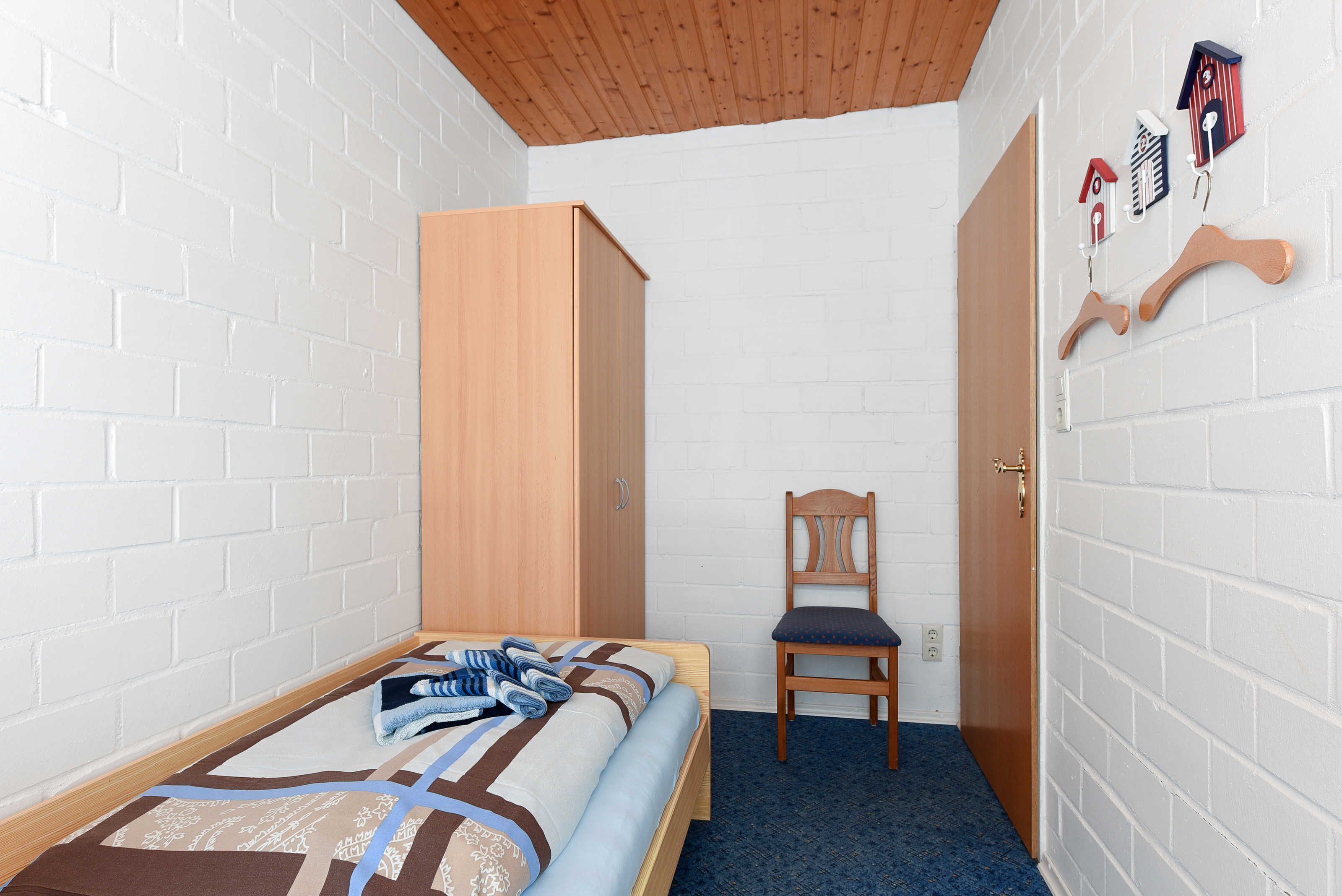 Ein kleines, ordentlich eingerichtetes Schlafzimmer mit einem Einzelbett, einem Holzschrank, einem Stuhl und einer Tür mit Griff.