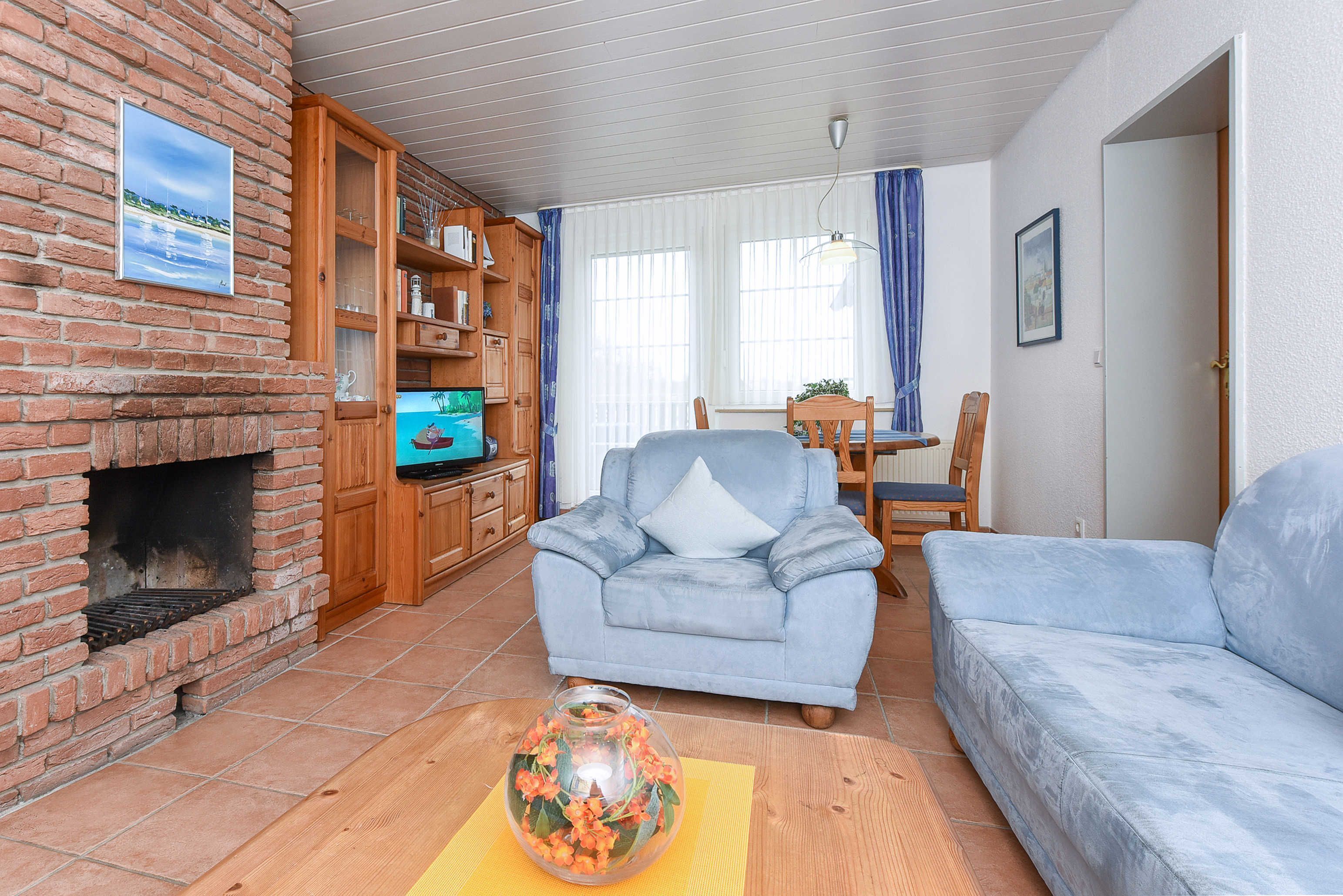 Ein Wohnzimmer mit Kamin aus Backsteinen, einem blauen Sessel, einer Couch, einem Holzschrank mit Büchern und einem Fernseher.