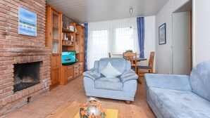 En stue med en murstenspejs, en blå lænestol, en sofa, et træskab med bøger og et fjernsyn.
