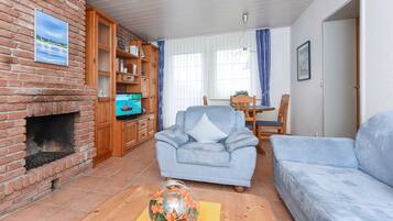 Ein Wohnzimmer mit Kamin aus Backsteinen, einem blauen Sessel, einer Couch, einem Holzschrank mit Büchern und einem Fernseher.