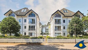 Un immeuble résidentiel moderne à deux étages, doté d’une façade symétrique, de grandes fenêtres et d’une clôture blanche en bois devant l’entrée.