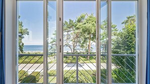 Une vue à travers une grande porte vitrée, agrémentée d’une rambarde en métal, qui donne sur un balcon d’où l’on peut admirer une plage et des arbres.