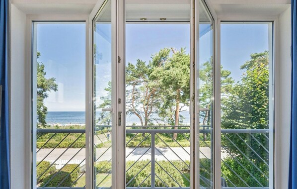 A view through a large glass door with a metal railing, leading to a balcony with a clear view of a beach and trees. - Wool Strandperle, white 25 - VS 25 (Heringsdorf)
