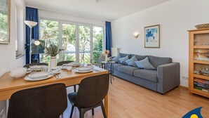 Un salon moderne avec un canapé gris, une table à manger dressée avec des assiettes et des verres, et une grande fenêtre donnant sur un paysage arboré.