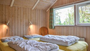 Ett rum med träväggar, ett stort fönster med utsikt över träd, två sängar med kuddar och två vägglampor.
