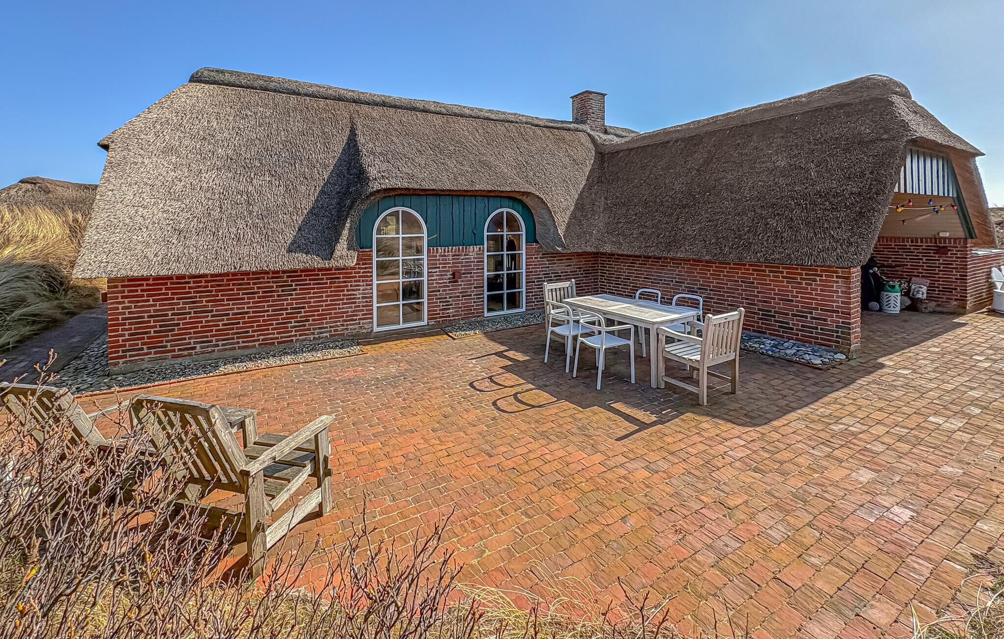 A thatched-roof house with a brick exterior, a wooden table and chairs, and a paved patio.