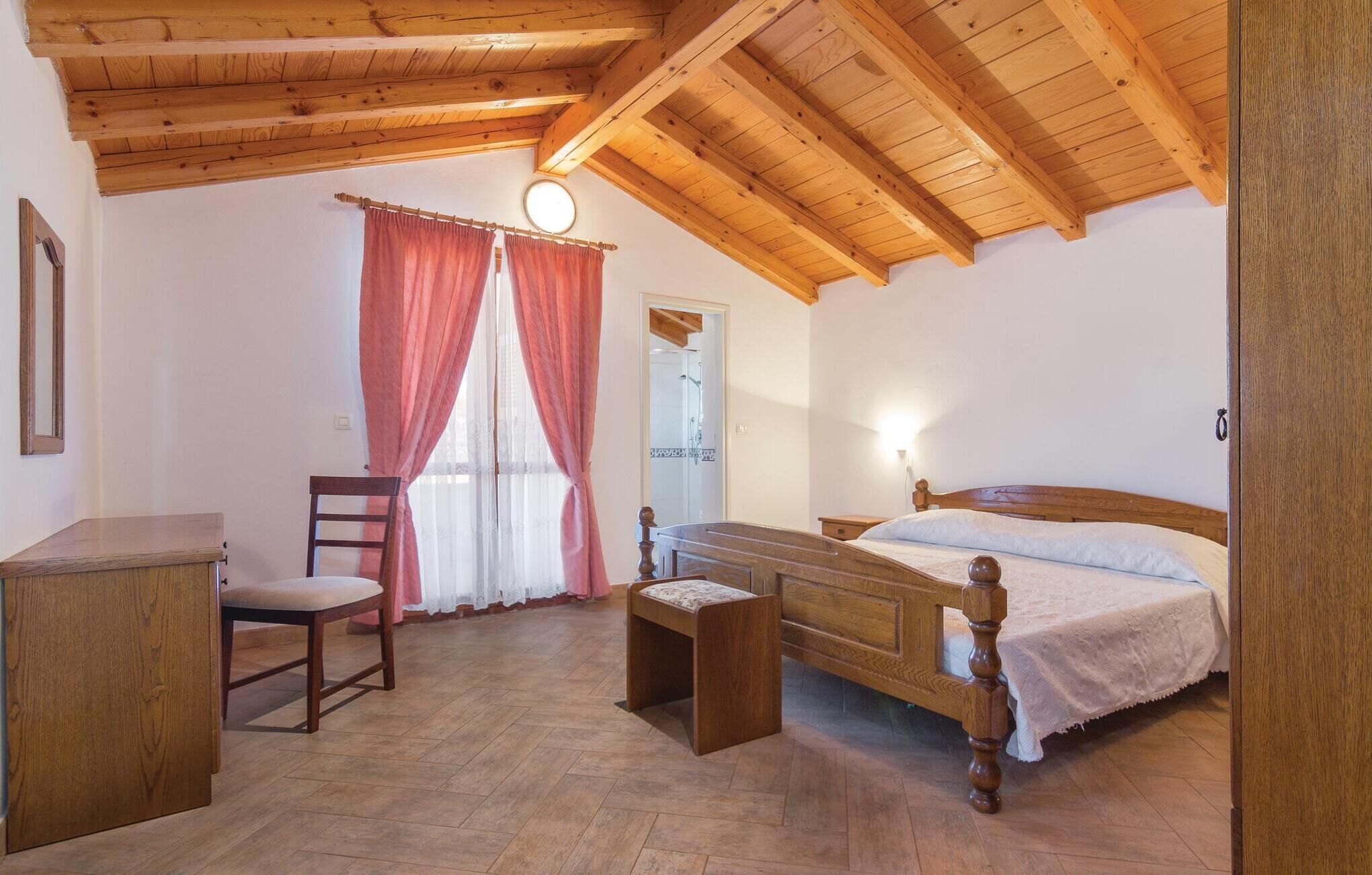 Une chambre avec un plafond en bois, un lit doté d’une tête de lit en bois, une chaise, une petite table et une fenêtre avec des rideaux.