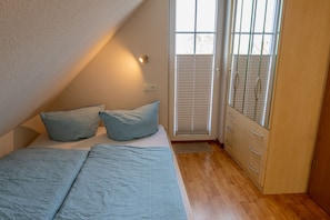 A bedroom with a bed, pillows, a dresser, and a window with blinds. - Vacation apartment 3 - Holiday apartments in Blu Hus (Kröslin)