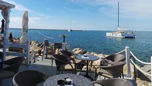 Une terrasse en bord de mer avec des sièges extérieurs, une vue sur la mer et un voilier à l’horizon.