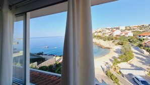 Une vue depuis une fenêtre donnant sur une zone côtière, avec des bâtiments, une plage et un ciel bleu dégagé.