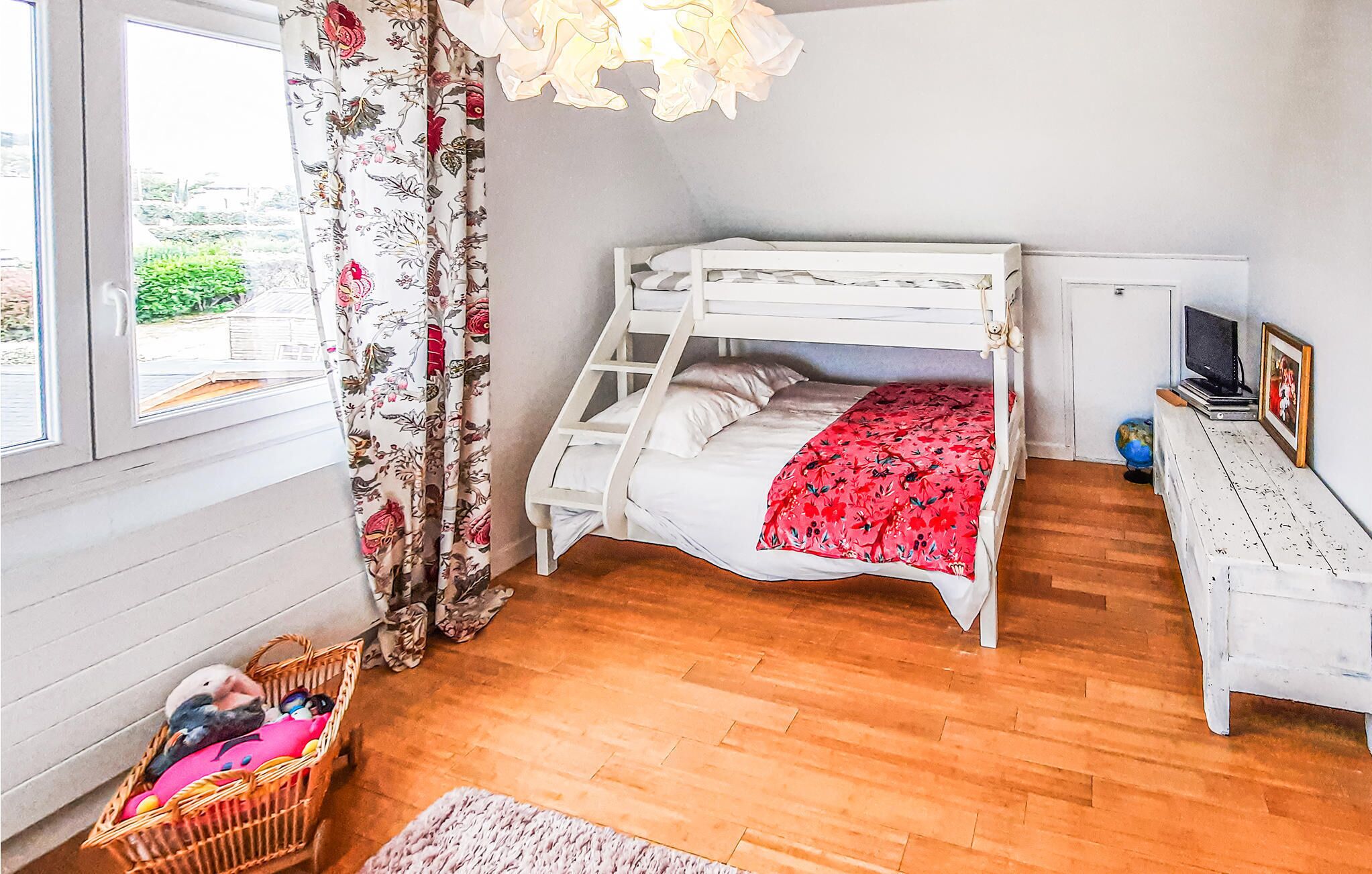 A bedroom with a bunk bed, floral curtains, a wicker basket with toys, a desk with a globe and picture frame, and a window with a view of greenery.