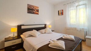 A hotel room with a bed, two bedside tables, a nightstand, a window with sheer curtains, and artwork on the walls.