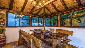 A rustic wooden dining area with a bench and table, large windows with a view of greenery, and a chandelier.
