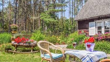 A rustic wooden cabin with a thatched roof, surrounded by a lush garden with various plants and flowers. There is a wicker chair and a small table with a checkered tablecloth and two wine glasses.