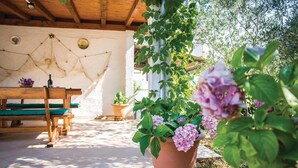 A wooden bench and a table with a potted plant and flowers in a sunny outdoor area with a white wall and wooden beams.