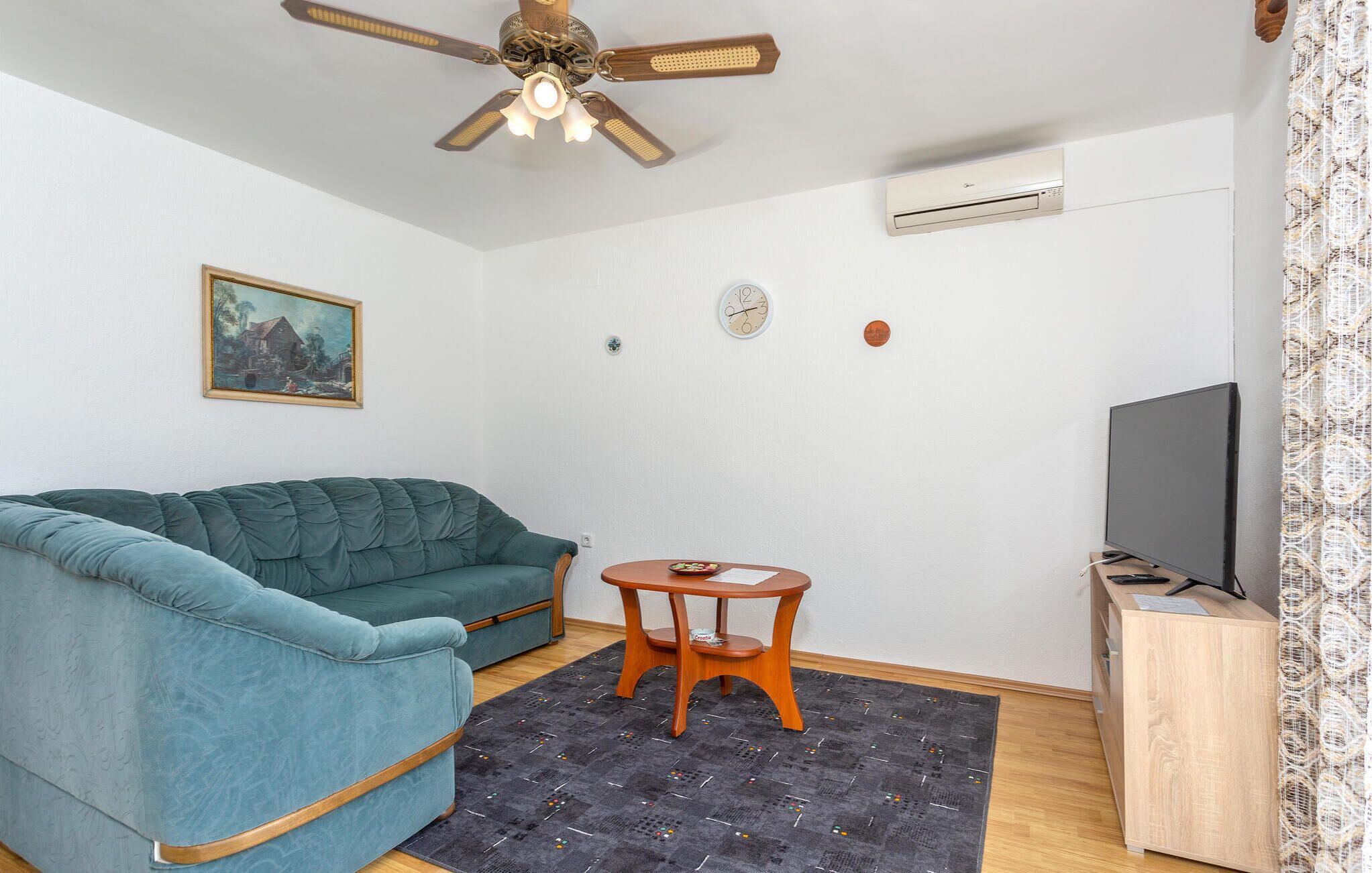 A living room with a teal sofa, a small wooden table, a ceiling fan, a wall-mounted air conditioner, a television, and a painting on the wall.