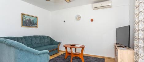 A living room with a teal sofa, a small wooden table, a ceiling fan, a wall-mounted air conditioner, a television, and a painting on the wall.