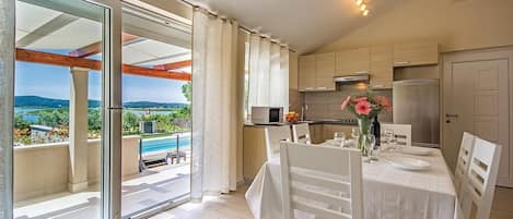 A dining area with a table set for four, a kitchen with modern appliances, and a view of a pool and landscape through large sliding glass doors.