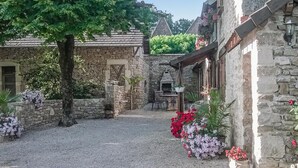 Une cour pavée de pierres, avec un chemin de gravier, entourée de bâtiments à l’architecture traditionnelle et agrémentée de plantes en pot.