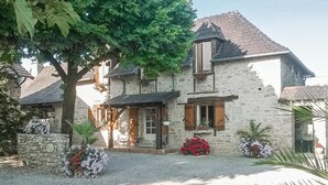 Une maison en pierre avec une allée gravillonnée, des volets en bois et un grand arbre devant.