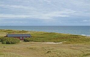 Ein Küstenhaus mit roter Fassade, umgeben von grasbewachsenen Dünen und Blick auf das Meer.