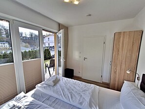 A bedroom with a bed, a wooden wardrobe, a door, and a window with a view of buildings and trees. - Villa Bettina 02 - Apartment 02 (Heringsdorf)