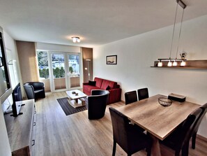 Een moderne woonkamer met een houten eettafel, zwarte stoelen, een roodgestreepte bank en een grote schuifpui naar een balkon.