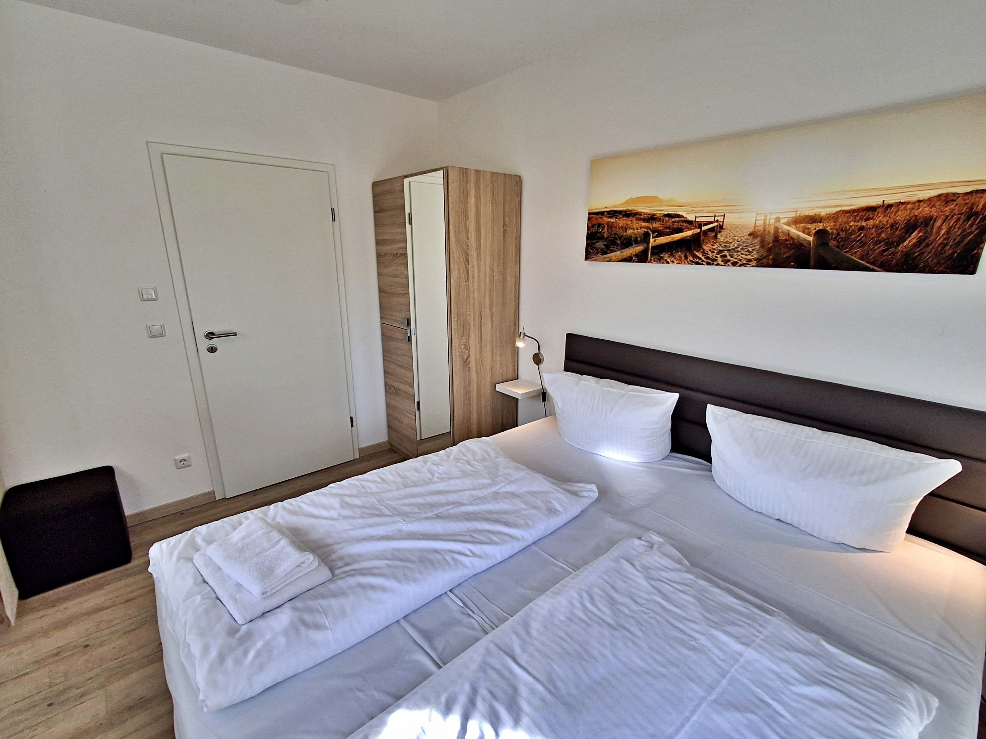 Ein ordentlich bezogenes Bett mit weißen Leinen und Kissen, ein hölzerner Kleiderschrank, ein kleiner Nachttisch mit Lampe und ein Wandbild mit Strandmotiv.