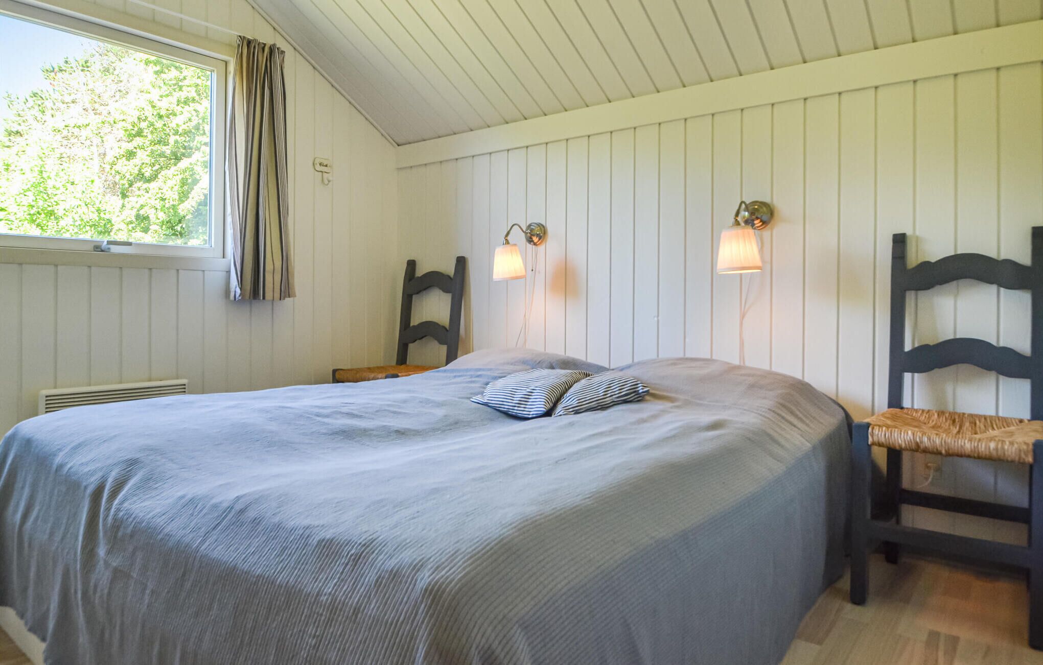Un dormitorio con cama, dos lámparas de pared, una silla y una ventana con cortinas.