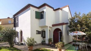 A two-story building with a white facade, a red door, and a small balcony. There is a patio area with a table and chairs under an umbrella. - Holiday apartment with air conditioning in a quiet location (Valbandon)